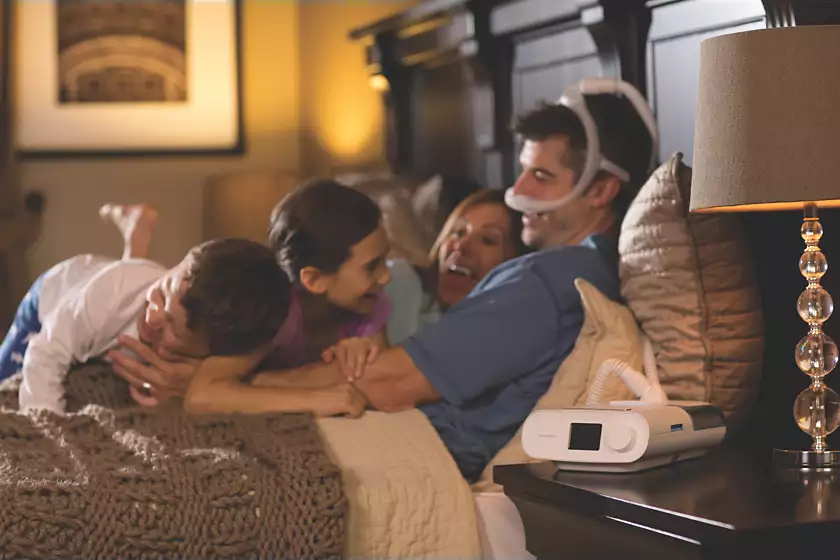 Philips Respironics DreamStation Auto CPAP and DreamWear Nasal Mask. A family of four in bed where the father has worn Philips Respironics DreamWear nasal mask and is using the Philips Respironics DreamStation Auto CPAP by the bedside.